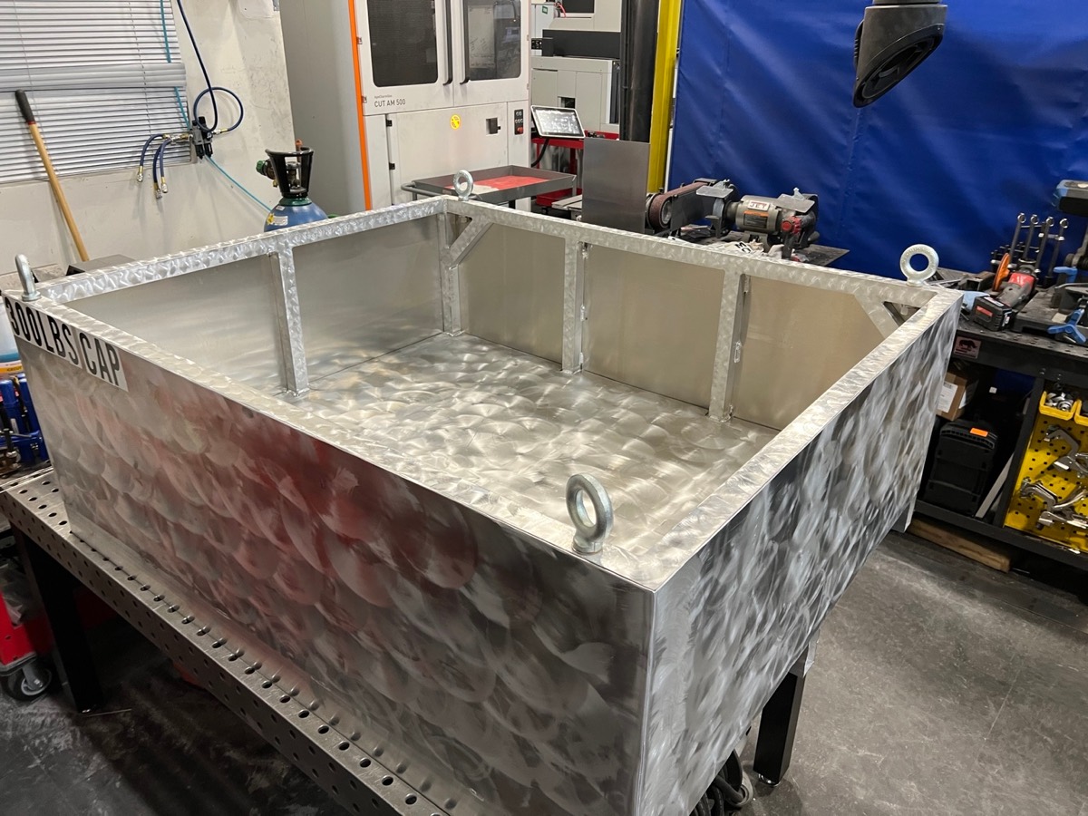 Fabricated metal tray with tall sides, reinforced corners, and a patterned metal finish in a workshop.