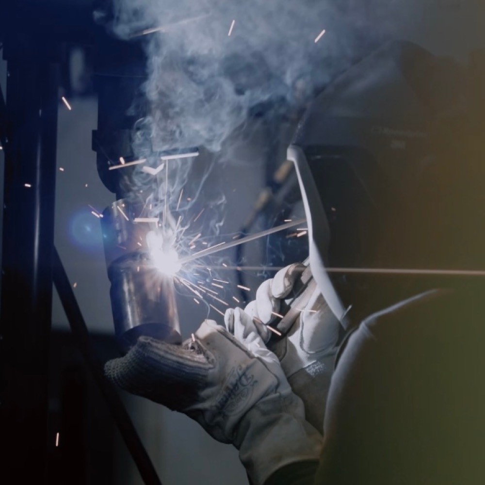 Welder in a protective helmet and gloves working on a metal joint, with sparks around the weld.